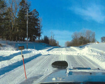 Snowplow looking down road with snow pushed across the road from a driveway