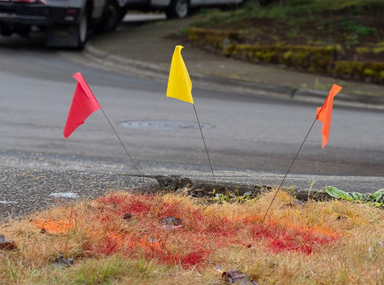 Underground utility marker flags on corner near asphalt road. Red on left, yellow in middle, and orange on right
