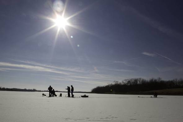 Four anglers stand on a snow- and ice-covered lake, silhouetted in front of a blue sky with the sun shining above them. 