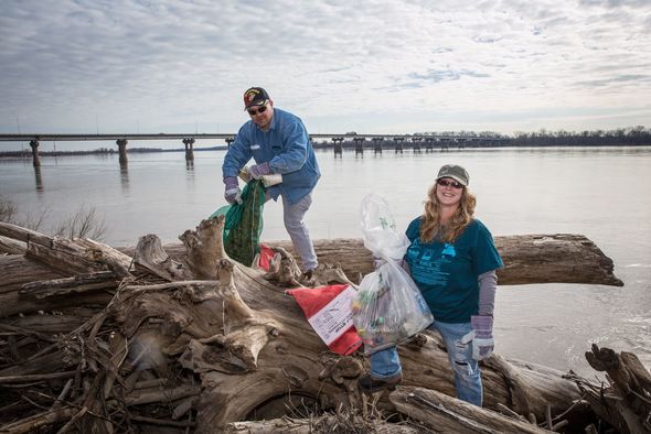 Two volunteers help clean trash along the Mississippi River with the Chain of Rocks Bridge in the background.
