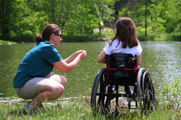 A mother helps her daughter in a wheelchair bait a hook in front of a lake.