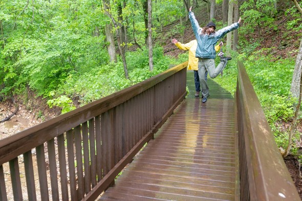 A female hiker does a jump with arms spread on a rain-soaked bridge over a creek.