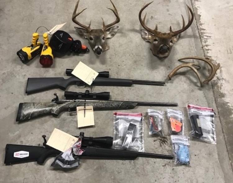 Several rifles, antlered deer heads, spotlights, and other poaching evidence in plastic bags displayed on a gray blanket on a concrete floor. 