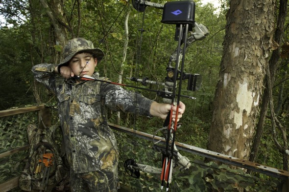 A male youth bowhunter dressed in camo takes aim with a longbow.
