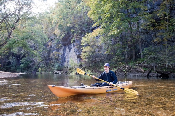 A young man paddles a kayak on the Huzzaah Creek with a bluff in the background.