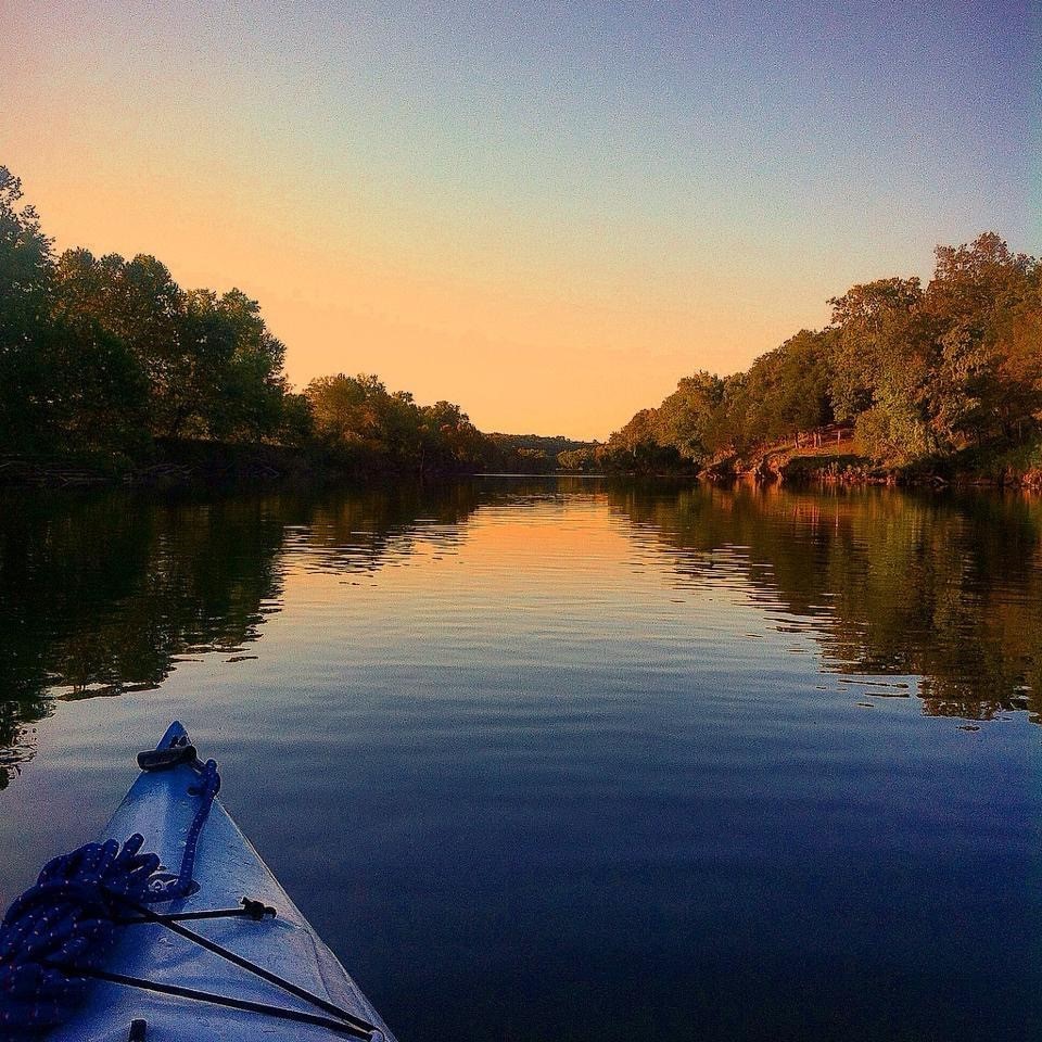 The bow of a blue kayak with blue coiled rope attached floats down a tree-lined river at sunset in early autumn. 