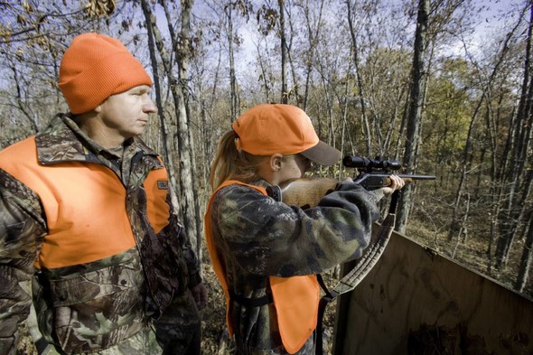 A young girl in camo and hunter orange vest and hat aims a rifle in the woods in autumn, her father stands behind her. 
