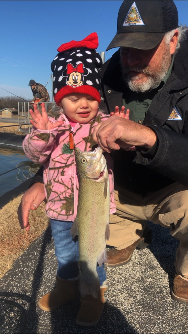 MDC staff member assisting a little girl holding a rainbow trout.