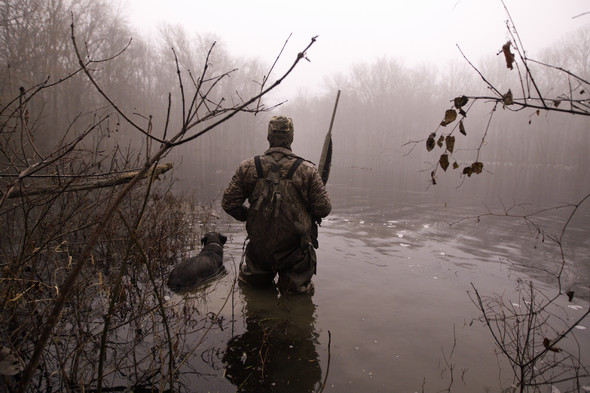 A lone male waterfowl hunter stands in the knee-deep waters of a foggy marsh with his hunting dog by his side.