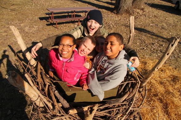 A group of young kids looks up at the camera while standing in a life size bald eagle nest replica.