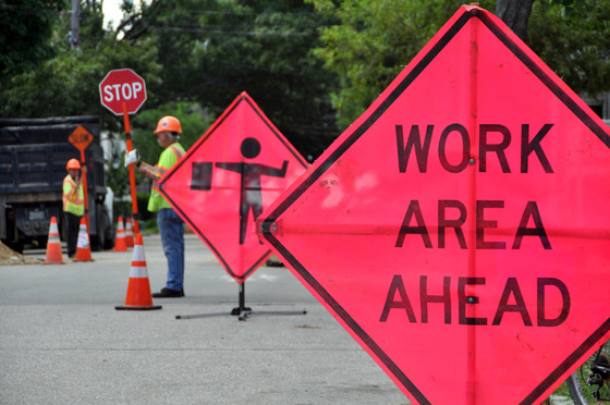 Construction zone sign in foreground says "Work Area Ahead" with workers in vests and construction cones in the background