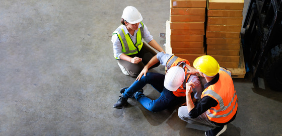 Two warehouse workers in safety vests and hard hats provide assistance to a colleague who has fallen on the concrete floor next to a stack of boxes.