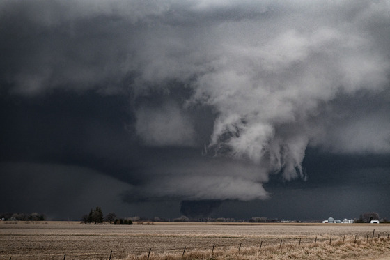 A massive, dark wall cloud and developing tornado loom over a flat, rural field under a heavy, grey sky.