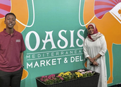 mother and son standing in front of their company wall that says Oasis Market and Deli