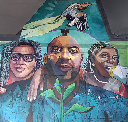 Three people. All have brown skin. One holds a megaphone; water flows from it. A heron flies. A sprout grows. The pattern of a shirt contains people.