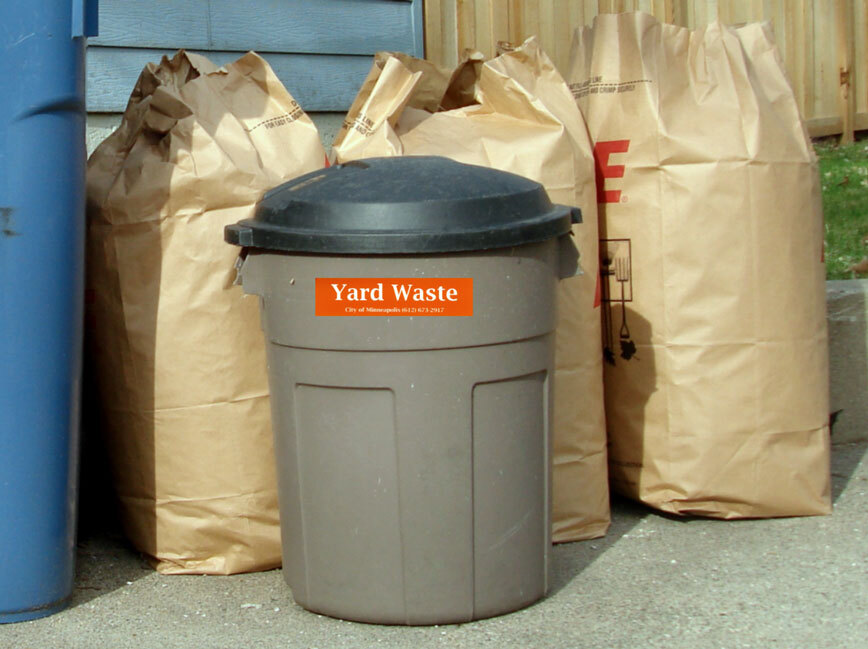 : Brown yard waste container with an orange “Yard Waste” sticker, placed in front of three large paper bags filled with yard waste