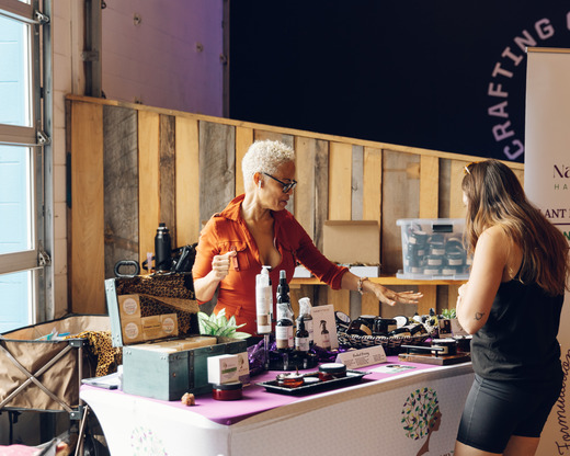 A woman stands behind a table at a market stall and talks with a customer during Black Business Week.