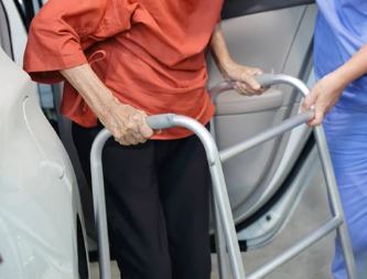 Elderly person using a walker to get out of a car with assistance from a person in blue scrubs