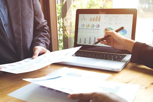 laptop screening with charts, sitting on a desk with two people arms holding papers