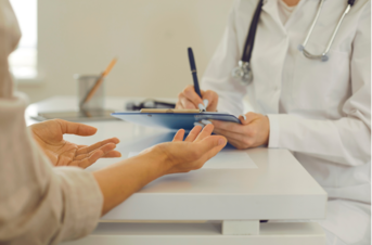 medical professional writing on clipboard with another person sitting at the table