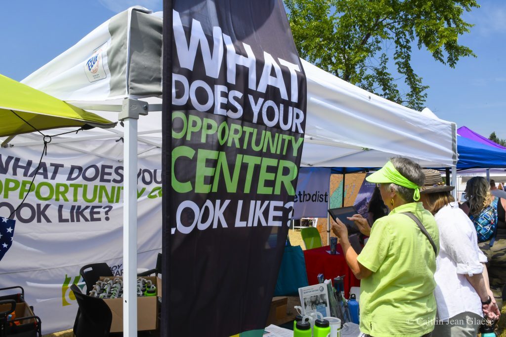 Two people are gathered around an outdoor tent with a sign that says "What does your opportunity center look like?"