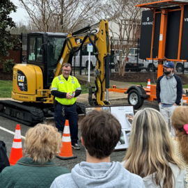 Person in yellow reflective shirt in front of a digger talking to a group of people