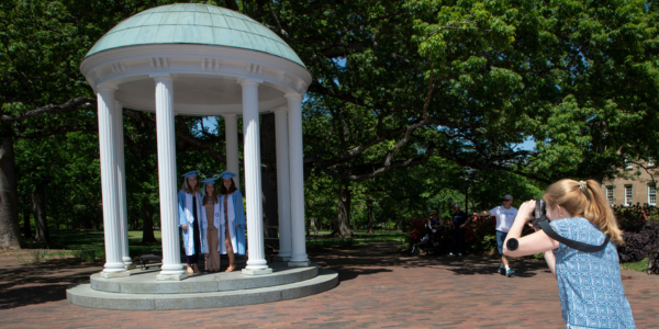 Person with pony tail taking picture of three UNC graduates in front of Old Well