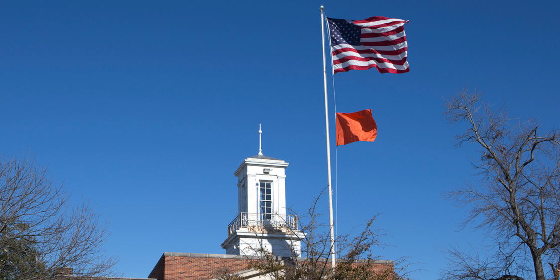 Orange flag and American flag on flag pole with blue sky and Post Office cupola in background