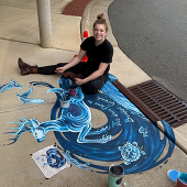 Artist sitting on sidewalk with mural of a creek and deer in blue
