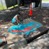 Artist on sidewalk painting nebulous blue and salmon shape at storm drain