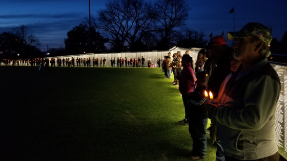 Image of a crowd of people standing at The Wall That Heals in the dark, honoring lost veterans.