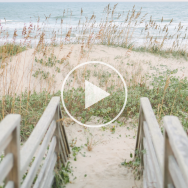 Image of a wooden walkway leading into the dunes along a beach. A "play video" button link sits over the image.