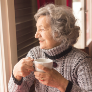 Image of a senior woman wearing a sweater and holding a tea cup, gazing out the window.
