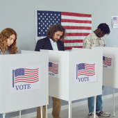 Image of one woman and two men voting using privacy voting boxes with U.S. flags on them.
