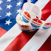 Image of a USA flag with a roll of "I Voted" stickers sitting on top.