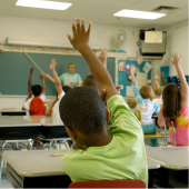 Image of a classroom of children with their hands raised.