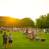 Image of people seated and standing on the lawn at Roanoke Island Festival Park watching a musical performance with drinks in hand.
