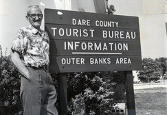 Image of Aycock Brown standing next to a sign that reads, "Dare County Tourist Bureau Information - Outer Banks Area"