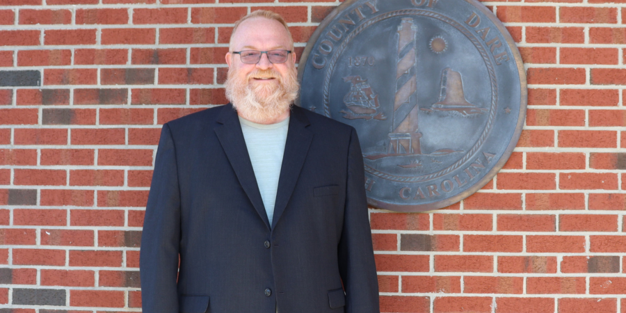 Image of Michael MacLean standing in front of the bronze Dare County seal.