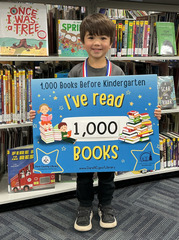 Smiling child holding "I read 1000 books before kindergarten sign" in front of new children's books at KDH Library