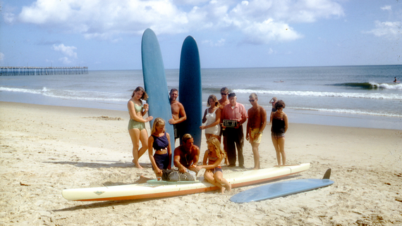 A group of men and women surfers stand together on the beach in front of a pier holding long surfboards in 1965.