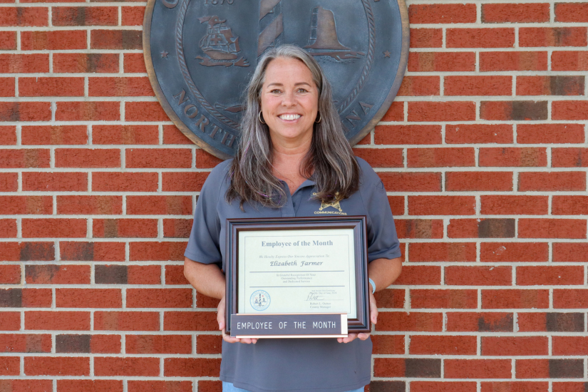 Dare County 911 Communications Center Telecommunicator Elizabeth Farmer stands in front of the bronze Dare County seal holding her framed certificate.