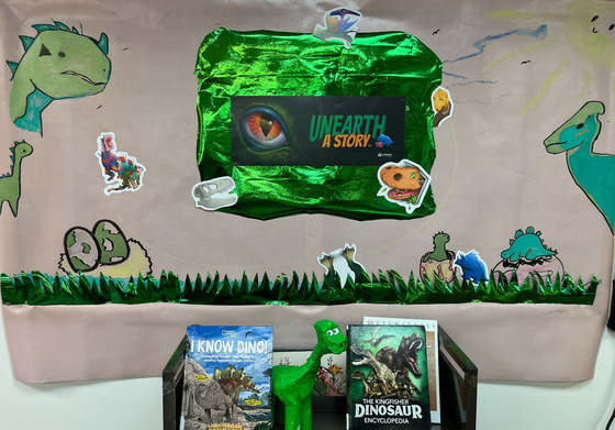 Unearth a Story bulletin board display for summer reading program with dinosaurs drawn by teen volunteer and related books displayed