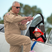 Image of a pilot standing on the wing demonstrating something to an audience while a boy in a toy fire hat sits in the cockpit.