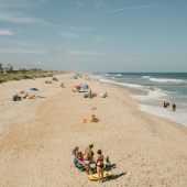 Image of people relaxing along the beach. Some sit under umbrellas, some in beach chairs and some stand in the shorebreak along the ocean.