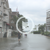 Image of a flooded downtown Manteo. Water fills the roadway and two boys and one adult wade through the waters.