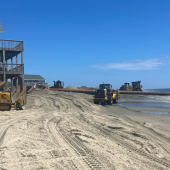 Image of machinery moving sand along the shore. A large pipe extends from the ocean to the shoreline. Several beach house sit along the shore.