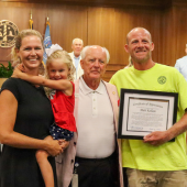 Image of Matt Kellam standing with his wife and daughter accepting the award from Chairman Woodard. 