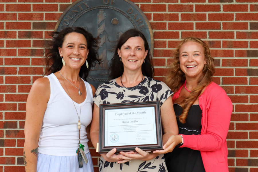 Jodi Wyant and Mandi Ferguson and stand beside Anna Miller in front of the Dare County seal after the presentation.