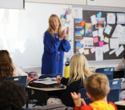 Image of a teacher clapping at the front of a classroom. Children at their desks clap with her.